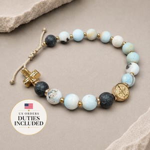Könnte beinhalten: Ein Perlenarmband mit hellblau-weiß marmorierten Steinen, dunkelgrauen, texturierten Perlen und kleinen goldfarbenen Zwischenelementen. Es verfügt über einen goldfarbenen Kreuzanhänger und eine runde Medaille mit verstellbarem beigem Kordelverschluss.