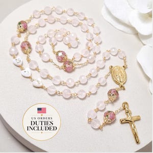 May include: A rosary featuring faceted pale pink beads, gold-tone metal links, and floral-patterned accent beads. It includes a gold-tone crucifix, a central medal depicting a figure, and three white heart-shaped beads with gold lettering.