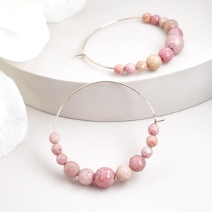 Puede incluir: Un par de pendientes de aro de oro con cuentas de piedras preciosas rosas y blancas. Los pendientes están sobre una superficie blanca.