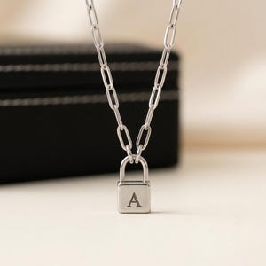 Op de afbeelding: Zilverkleurige ketting met een hangslot-hanger met de letter "A". De hanger hangt aan een fijne ketting met langwerpige schakels. De achtergrond toont een zwarte doos en een neutraal oppervlak.