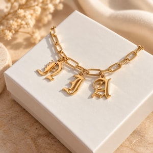 May include: A gold-toned chain bracelet featuring three dangling letter charms in an ornate, gothic-style font. The bracelet is constructed with elongated rectangular links and is displayed on a plain white square box.