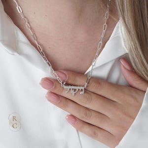 May include: A silver-toned necklace featuring a paperclip-style chain and a central pendant displaying the name Nicole written in Hebrew script. The metallic finish is polished and reflective, creating a clean and bright appearance against the white fabric.