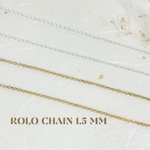 Puede incluir: Primer plano de tres delicadas cadenas Rolo en tonos plateados y dorados. Las cadenas tienen 1,5 mm de grosor y se muestran sobre un fondo texturizado blanquecino. El texto "ROLO CHAIN 1.5 MM" está impreso en la parte inferior de la imagen.