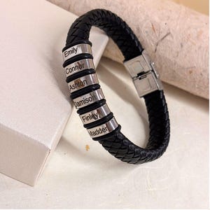 May include: A black braided leather bracelet featuring six cylindrical silver-toned metal beads. Each bead is engraved with a name: Emily, Connor, Ashton, Jamison, Finley, and Madder. The bracelet includes a silver-toned metal clasp closure.