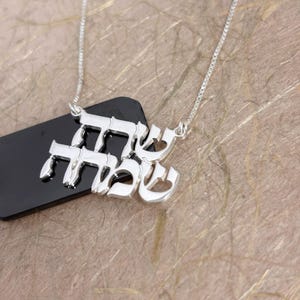 May include: A silver necklace featuring a pendant with Hebrew script characters. The pendant is suspended from a delicate silver box chain. The metallic surface of the script is polished and reflective, showcasing the intricate letter forms clearly.