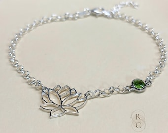 May include: A silver chain bracelet featuring a delicate lotus flower charm and a single green gemstone. The bracelet is displayed on a beige surface.