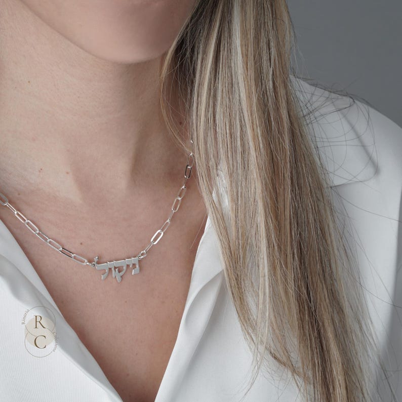 May include: A silver-toned necklace featuring a polished metal nameplate with Hebrew script lettering. The pendant is suspended from a delicate chain composed of elongated, oval-shaped links, creating a clean and modern aesthetic for the jewelry piece.