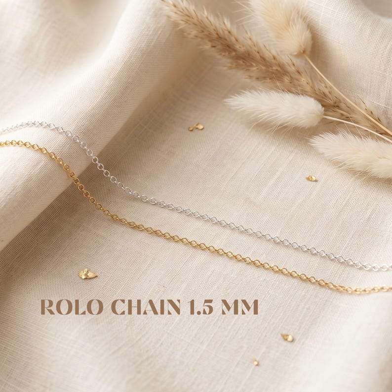 May include: Two rolo chains, one in a silver tone and one in a gold tone, each measuring 1.5 mm in width. The text ROLO CHAIN 1.5 MM is printed in brown letters below the chains, which are draped across a textured, off-white fabric surface.