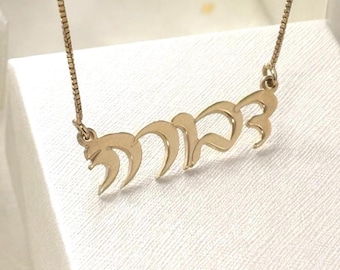 May include: A gold-toned necklace featuring a pendant with Hebrew script lettering. The pendant is suspended from a delicate box chain. The metal has a polished finish, and the script is connected in a single, continuous piece of metalwork.