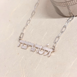 May include: A silver-toned necklace featuring a polished metal nameplate with Hebrew script lettering. The pendant is attached to a delicate paperclip-style chain, creating a cohesive metallic look with a bright, reflective finish throughout the piece.
