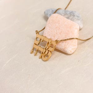 May include: A gold-toned necklace featuring a pendant with Hebrew characters arranged in a stacked, block-like formation. The pendant is attached to a delicate box chain, showcasing a polished metallic finish and intricate letter cutouts.