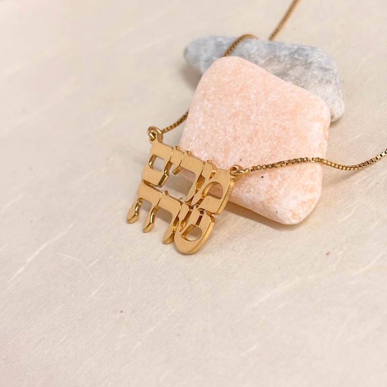 May include: A gold-toned necklace featuring a pendant with Hebrew characters arranged in a stacked, block-like formation. The pendant is attached to a delicate box chain, showcasing a polished metallic finish and intricate letter cutouts.