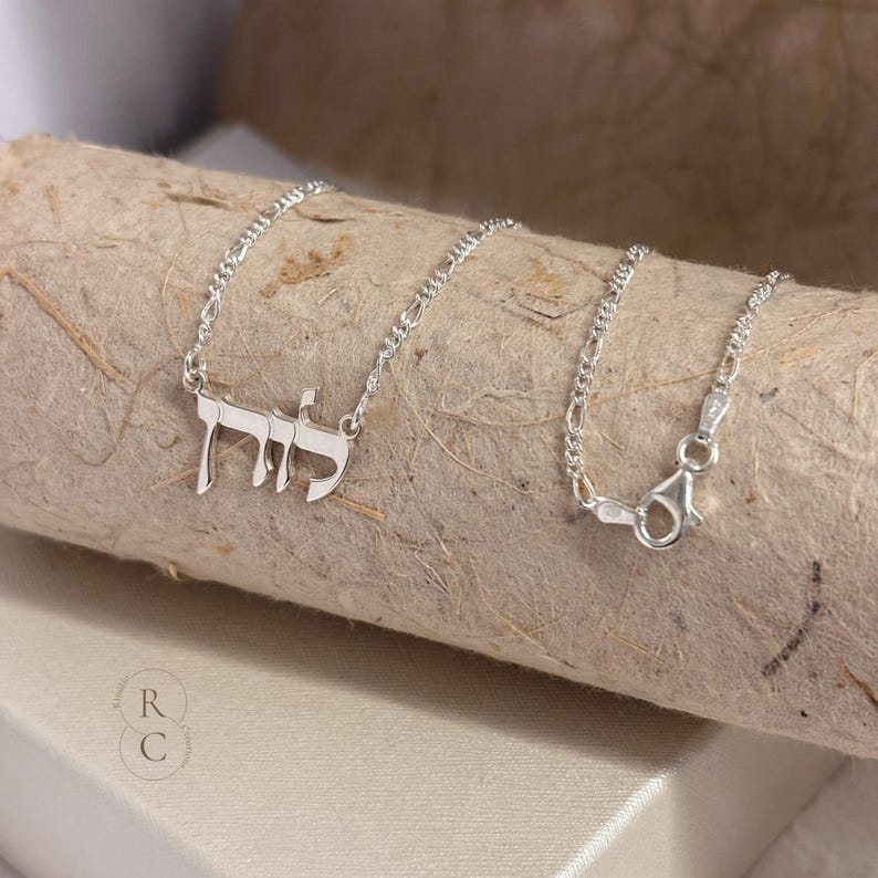 May include: A silver necklace featuring a nameplate pendant with Hebrew script characters. The pendant is attached to a delicate silver chain with a lobster claw clasp. The metal has a polished, reflective finish, and the chain links are oval.