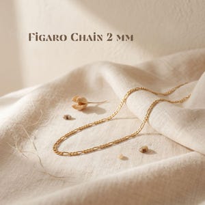 May include: A gold-toned Figaro chain necklace with a 2 mm width, as indicated by the text 'FIGARO CHAIN 2 MM' displayed above the jewelry. The chain features a repeating pattern of elongated oval links interspersed with smaller circular links.