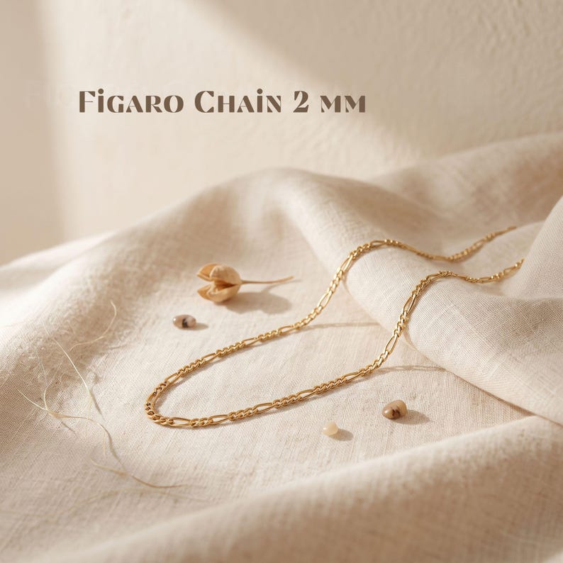 May include: A gold-toned Figaro chain necklace with a 2 mm width, as indicated by the text 'FIGARO CHAIN 2 MM' displayed above the jewelry. The chain features a repeating pattern of elongated oval links interspersed with smaller circular links.