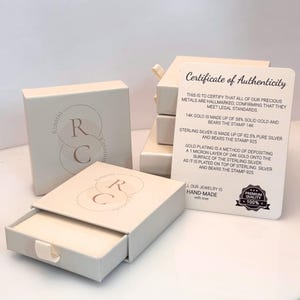 May include: Off-white square jewelry boxes with a slide-out drawer featuring a circular logo with the letters RC and the text Rinatia Creation. A white Certificate of Authenticity card details gold and silver standards and includes a 100% quality seal.