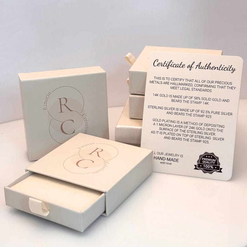 May include: Off-white square jewelry boxes with a slide-out drawer featuring a circular logo with the letters RC and the text Rinatia Creation. A white Certificate of Authenticity card details gold and silver standards and includes a 100% quality seal.