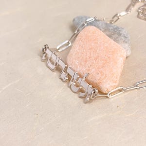 May include: A silver-toned metal necklace featuring a nameplate pendant with Hebrew script lettering. The pendant is attached to a delicate paperclip-style chain. The jewelry piece is positioned resting against a smooth, light-colored, rectangular stone.