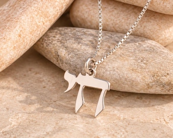 Può includere: Una collana color argento con un ciondolo a forma del carattere ebraico Chai. Il ciondolo è sospeso a una sottile catena a maglie quadrate. Il metallo ha una finitura lucida e riflettente, e il design è composto da due lettere ebraiche.