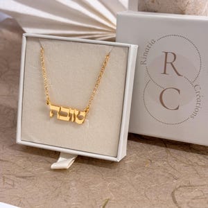 May include: A gold-colored necklace featuring a nameplate pendant with Hebrew script lettering. The necklace is displayed inside a white square gift box that has the text Rinatia Cr&eacute;ations and the letters R and C printed on the lid.