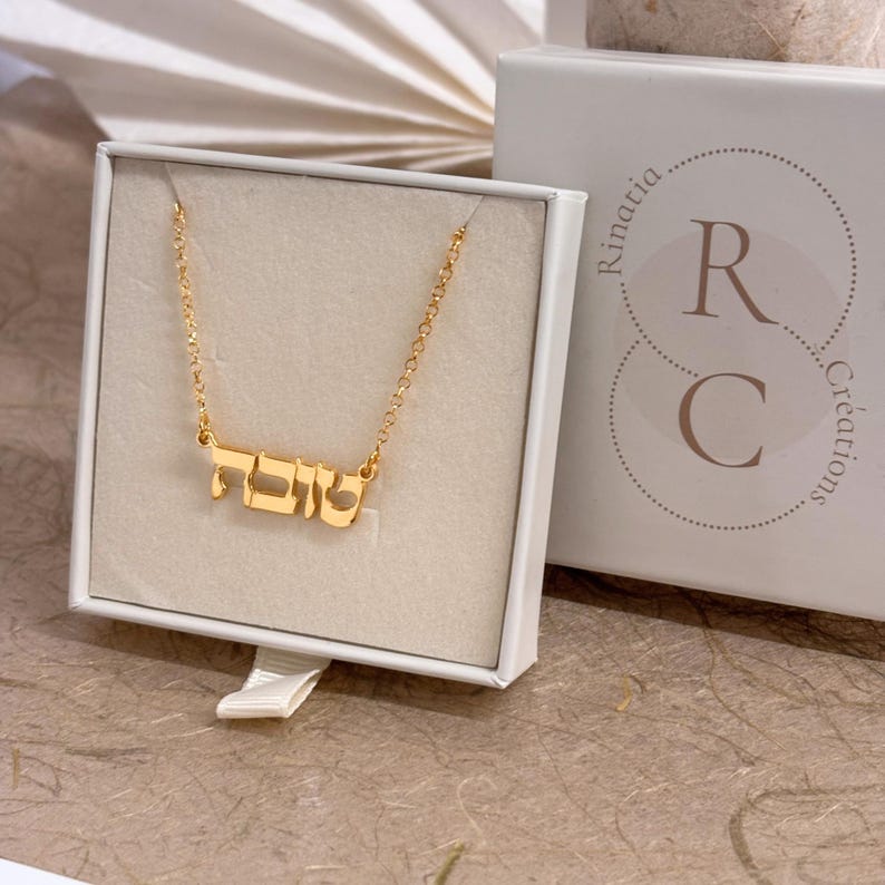May include: A gold-colored necklace featuring a nameplate pendant with Hebrew script lettering. The necklace is displayed inside a white square gift box that has the text Rinatia Cr&eacute;ations and the letters R and C printed on the lid.