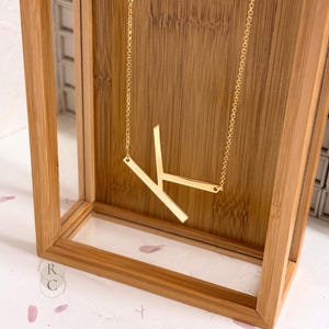 May include: A gold-coloured necklace featuring a large, stylised capital letter K pendant suspended from a delicate chain. The pendant is constructed from flat, polished metal bars that form the shape of the letter, creating a clean, geometric look.
