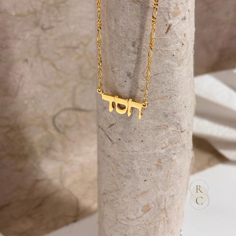 May include: A gold-toned necklace featuring a delicate chain and a pendant with Hebrew lettering. The pendant displays the word 'Chesed' in a polished, metallic finish, suspended from a fine link chain that catches the light.