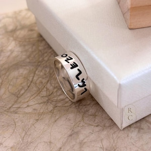 May include: A silver-toned metal ring with a smooth, polished finish. The outer surface of the band features black engraved Hebrew lettering that wraps around the circumference of the ring, creating a distinct contrast against the metallic surface.