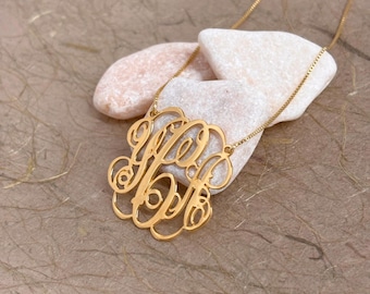 Puede incluir: Un collar de tono dorado con un colgante circular que presenta un diseño de monograma intrincado e interconectado. El colgante cuelga de una cadena fina y delicada, mostrando un acabado metálico pulido y letras ornamentadas en cursiva.