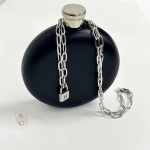 Może przedstawiać: Czarna flaszka z srebrnym łańcuszkiem i kłódką. Kłódka ma wygrawerowaną literę "H".