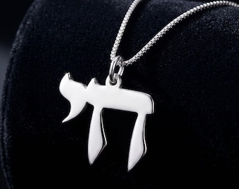 Può includere: Una collana color argento lucido con un ciondolo a forma di lettera ebraica Chai. Il ciondolo è sospeso a una delicata catenina a maglie quadrate, caratterizzata da una superficie metallica liscia e riflettente con bordi netti e definiti.