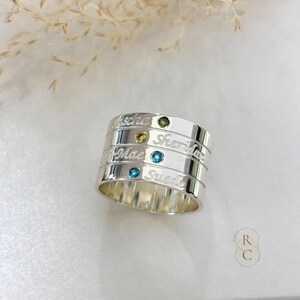 Puede incluir: Un conjunto de anillos apilables en tono plateado compuesto por cuatro bandas individuales. Cada banda presenta un nombre grabado, incluyendo Jascha, Sheridan, Mae y Suede, acentuado con pequeñas piedras preciosas redondas incrustadas en tonos azules y verdes.