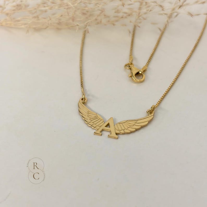 Pu&ograve; includere: Collana in oro con un ciondolo a forma di lettera A e ali. La collana &egrave; su uno sfondo bianco.