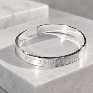 May include: A polished silver cuff bracelet with an open design. The outer surface of the metal band is engraved with the geographic coordinates 49° 25' 14.8" N. The bracelet features a smooth, reflective finish and a simple, circular shape.
