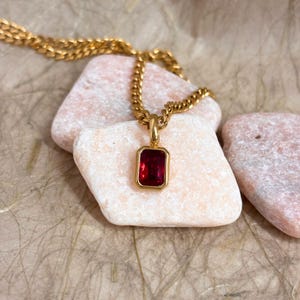 May include: A gold-toned chain necklace featuring a rectangular pendant with a vibrant red gemstone set in a smooth gold-colored bezel. The pendant hangs from a delicate link chain, showcasing a polished finish and a classic, clean design.