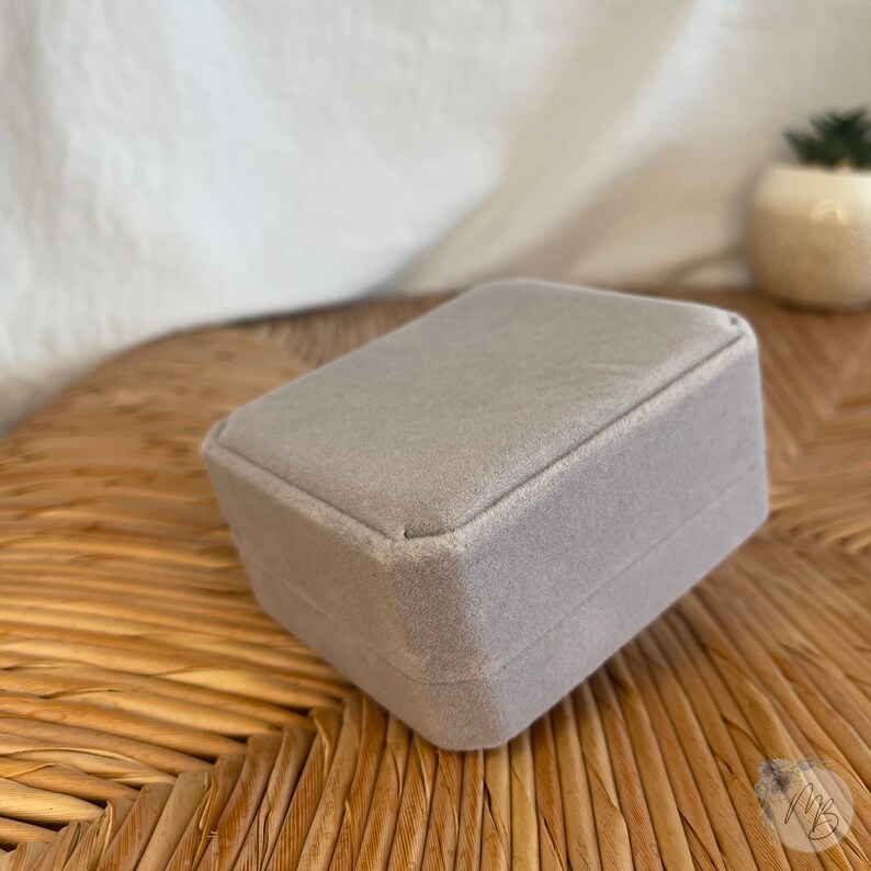 Peut inclure: Une bo&icirc;te &agrave; bijoux en velours gris de forme carr&eacute;e. La bo&icirc;te est pos&eacute;e sur une surface tiss&eacute;e.