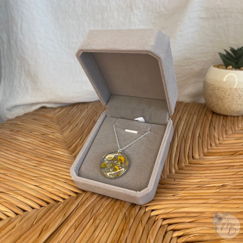 Peut inclure: Un collier en cha&icirc;ne argent avec un pendentif rond pr&eacute;sentant des fleurs jaunes et blanches press&eacute;es enrob&eacute;es de r&eacute;sine transparente. Le collier est pr&eacute;sent&eacute; dans un &eacute;crin en velours gris.