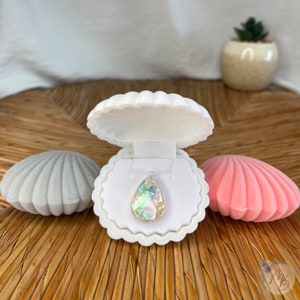 May include: A teardrop-shaped iridescent pendant necklace in a white seashell-shaped jewellery box. The box is open and the necklace is inside. There are two other seashell-shaped boxes, one grey and one pink, on either side of the white box.