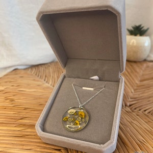 Peut inclure: Un collier en cha&icirc;ne argent avec un pendentif rond pr&eacute;sentant des fleurs jaunes et blanches press&eacute;es ench&acirc;ss&eacute;es dans de la r&eacute;sine transparente.