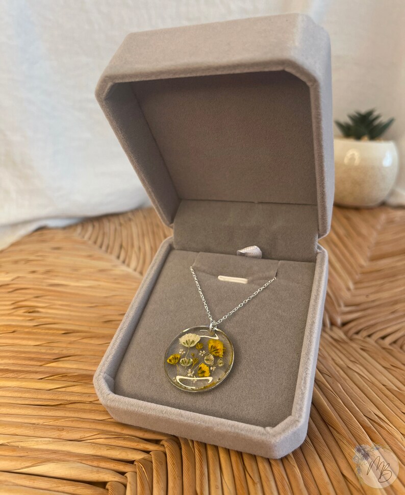 Peut inclure: Un collier en cha&icirc;ne argent avec un pendentif rond pr&eacute;sentant des fleurs jaunes et blanches press&eacute;es ench&acirc;ss&eacute;es dans de la r&eacute;sine transparente.