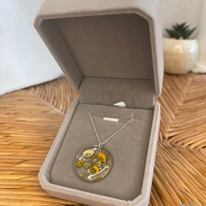 Peut inclure: Un collier en cha&icirc;ne argent avec un pendentif rond pr&eacute;sentant des fleurs jaunes et blanches press&eacute;es ench&acirc;ss&eacute;es dans de la r&eacute;sine transparente. Le collier est pr&eacute;sent&eacute; dans un &eacute;crin en velours gris.