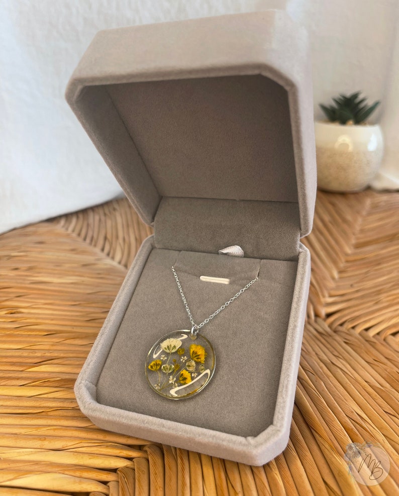 Peut inclure: Un collier en cha&icirc;ne argent avec un pendentif rond pr&eacute;sentant des fleurs jaunes et blanches press&eacute;es ench&acirc;ss&eacute;es dans de la r&eacute;sine transparente. Le collier est pr&eacute;sent&eacute; dans un &eacute;crin en velours gris.