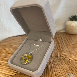 Peut inclure: Un collier en cha&icirc;ne argent avec un pendentif rond pr&eacute;sentant des fleurs s&eacute;ch&eacute;es press&eacute;es dans des tons de jaune et de blanc ench&acirc;ss&eacute;es dans de la r&eacute;sine transparente. Le collier est pr&eacute;sent&eacute; dans un &eacute;crin en velours gris.