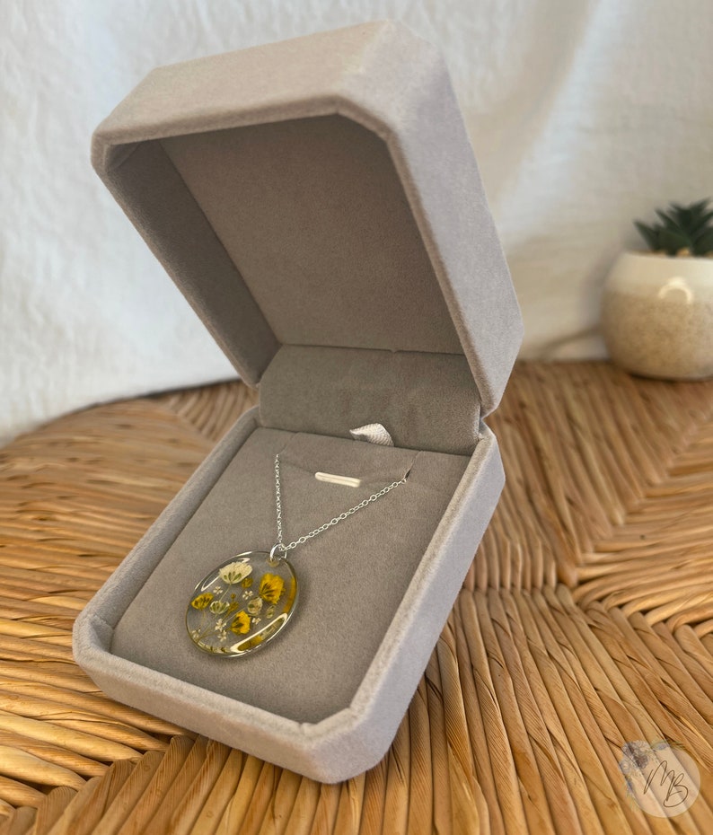 Peut inclure: Un collier en cha&icirc;ne argent avec un pendentif rond pr&eacute;sentant des fleurs s&eacute;ch&eacute;es press&eacute;es dans des tons de jaune et de blanc ench&acirc;ss&eacute;es dans de la r&eacute;sine transparente. Le collier est pr&eacute;sent&eacute; dans un &eacute;crin en velours gris.