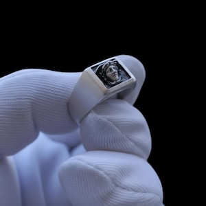 May include: A silver-coloured signet ring featuring a rectangular face with a raised, detailed relief of a classical human head profile. The metal surface has a brushed finish, and the recessed areas around the central figure are darkened for contrast.