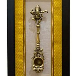 Puede incluir: Una escultura de bronce enmarcada de una figura de Ganesha bailando sobre un soporte decorativo. El marco tiene un borde interior dorado y un borde exterior amarillo con un patrón texturizado.