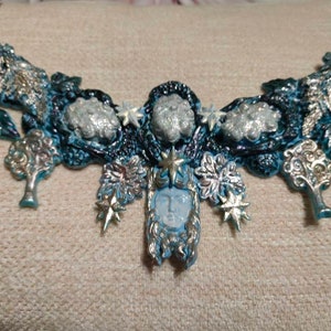 May include: A blue and silver necklace with a central pendant featuring a face surrounded by leaves and stars. The necklace is made of polymer clay and has a textured surface.