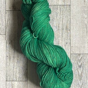 May include: A skein of green yarn, wound into a ball shape, on a light wood background.