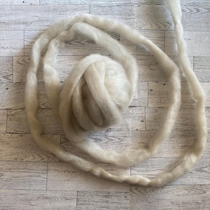 May include: A ball of off-white roving yarn with a long strand extending outward. The yarn is soft and fluffy, ideal for various fiber art projects. The yarn is displayed on a light wood grain surface.
