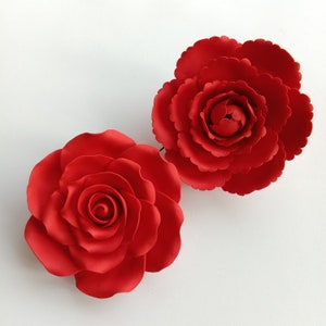 May include: Two bright red artificial flowers, a rose and a peony, are set against a white backdrop. The rose has tightly wound petals, while the peony has a layered, ruffled appearance. These decorative flowers are suitable for crafting or home decor.
